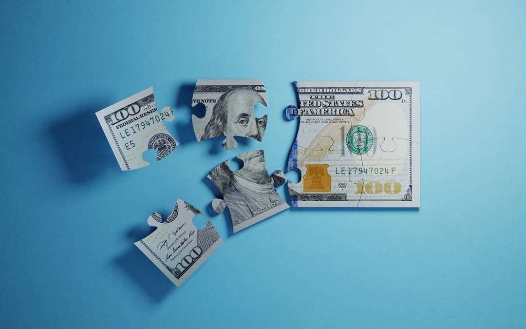 A U.S. hundred-dollar bill cut into jigsaw pieces on a blue background, only partly put together.