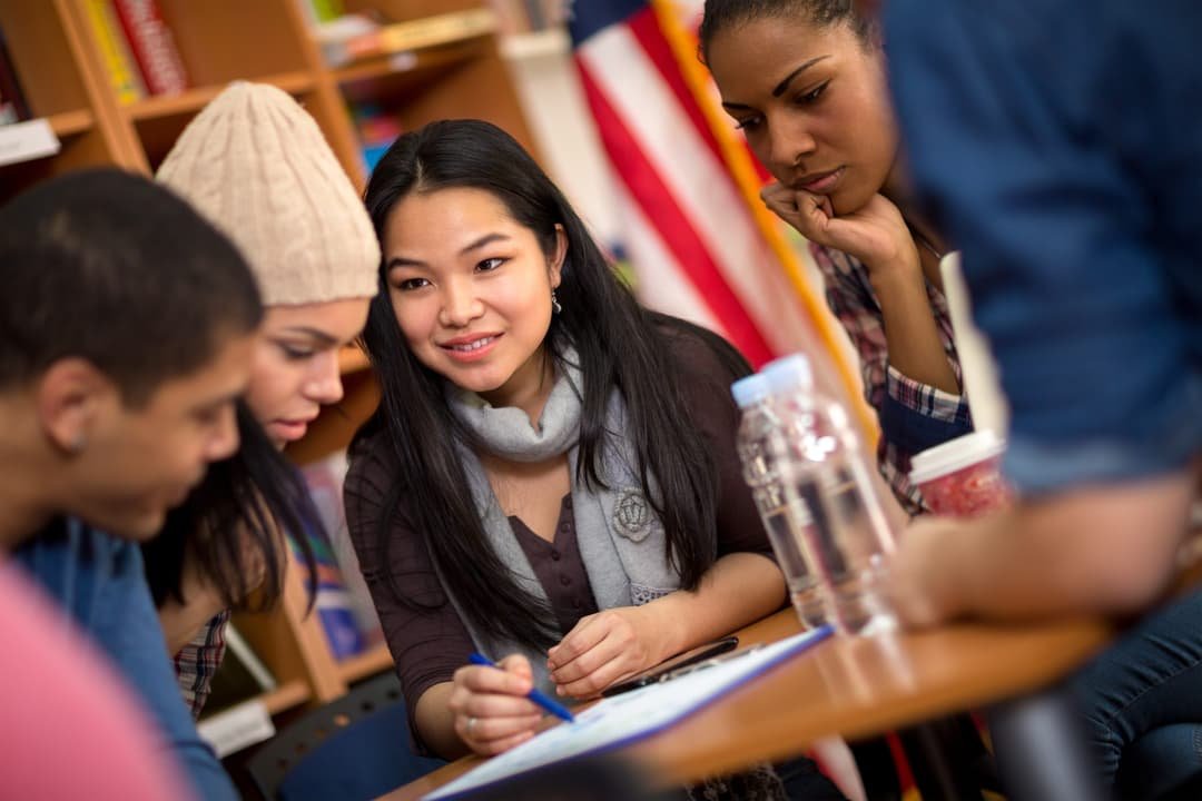 Students around a table working through a document together, with a U.S. flag in the background.